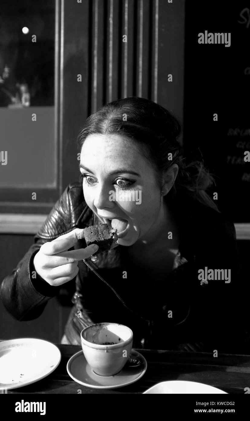 Giovane donna nel suo 20s a bere caffè cappuccino e mangiare un brownie al cioccolato al di fuori di un cafe nelle corsie in area di Brighton Regno Unito Foto Stock