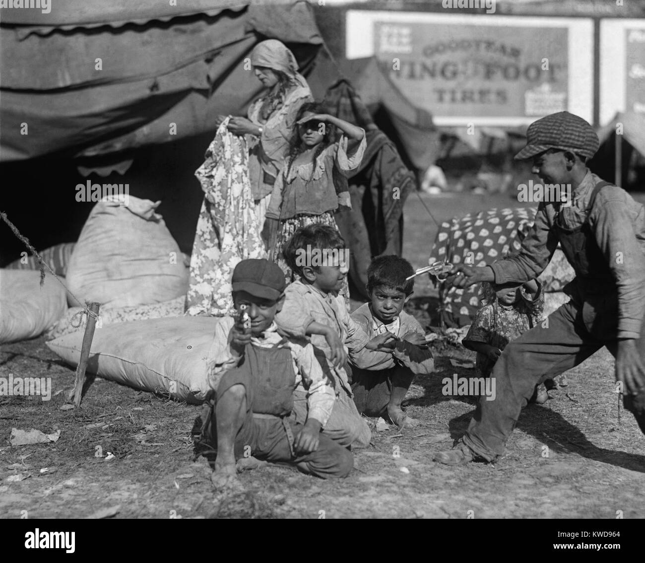 Gli zingari bambini a giocare in un accampamento, eventualmente un circo, in Washington, D.C. Vicinanze, 1923. Vicino a una donna che tiene un indumento fra diversi sacchetti ripieni in una tenda. In fondo è un cartellone per Goodyear, "Ala piede" pneumatici (BSLOC 2016 8 82) Foto Stock