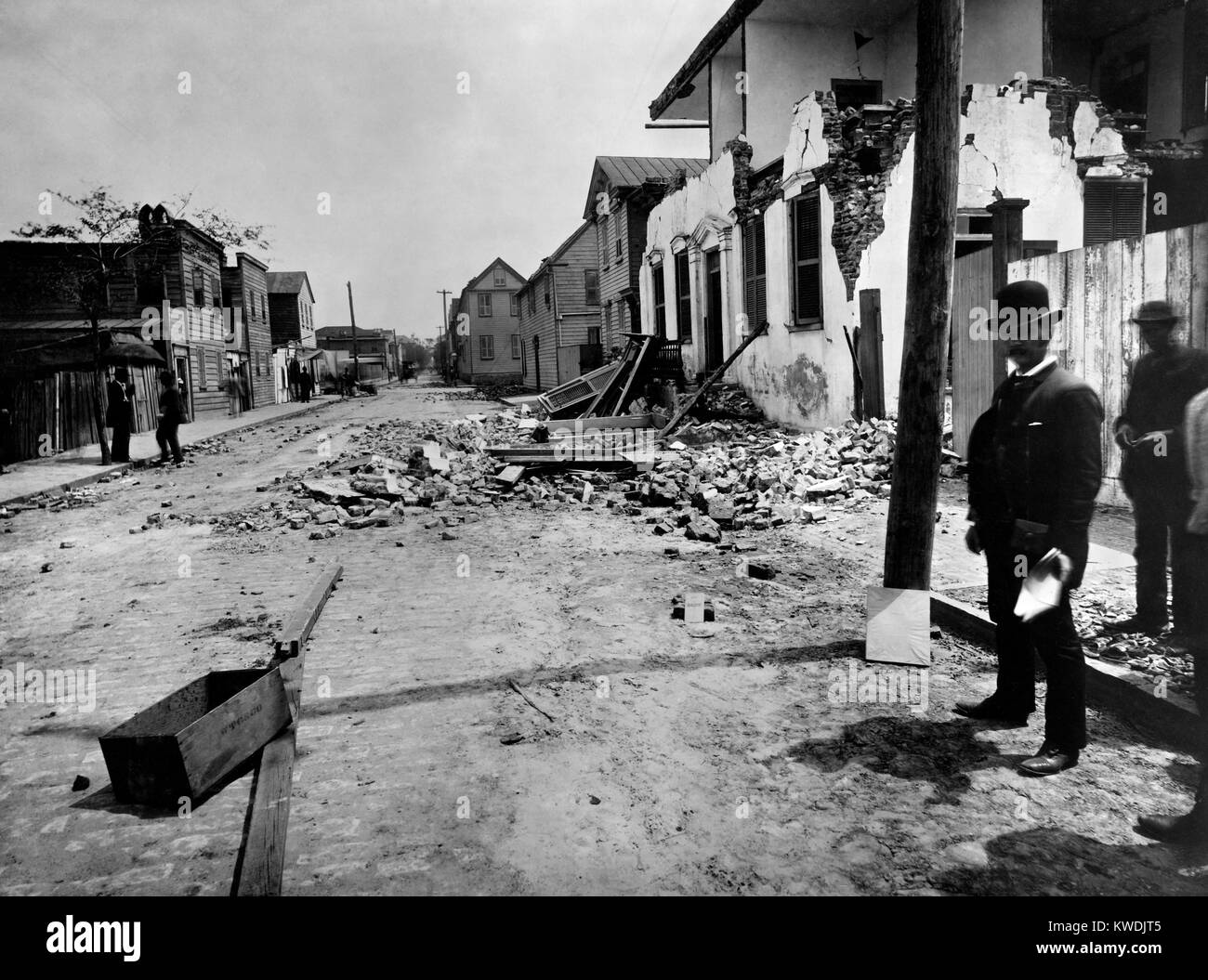 Casa di mattoni su Tradd Street, Charleston, danneggiate dal terremoto del mese di agosto 31, 1886. William John McGee dell'U.S. Geological Survey sorge nei pressi di un marciapiede fessura che è coperta da due tavole. Un cartone focimeter è puntellato contro il polo utilità, all'aiuto per la messa a fuoco della fotocamera. Foto di John K. Hillers (BSLOC 2017 17 56) Foto Stock