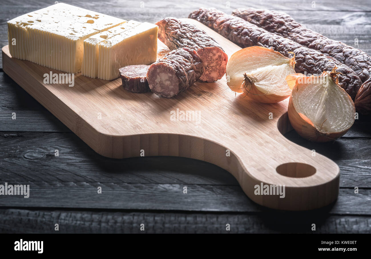 Il gustoso pasto con fatti in casa affumicate salsicce di carne di manzo e di maiale, formaggio fresco e cipolla, visualizzata su un tagliere di legno su un rustico tavolo nero. Foto Stock