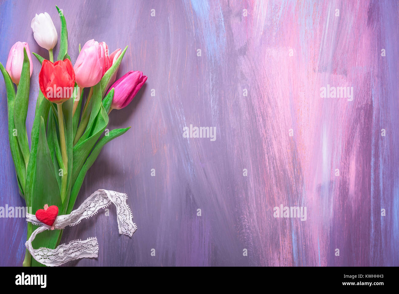 Bel bouquet di tulipani colorati legati con nastro in pizzo e un clip in legno con cuore rosso visualizzato su uno sfondo di legno con molte sfumature di viola. Foto Stock