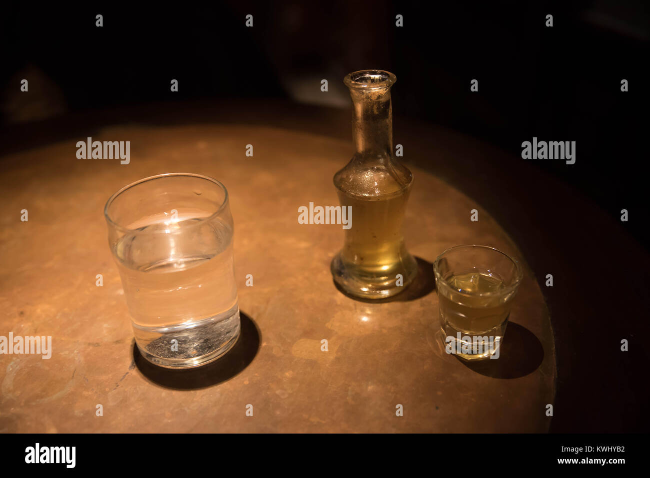 A snap shot e una bottiglia riempita con tradizionale greco (rakomelo raki con miele) accanto ad un bicchiere di acqua in un tavolo di legno con luce scarsa Foto Stock