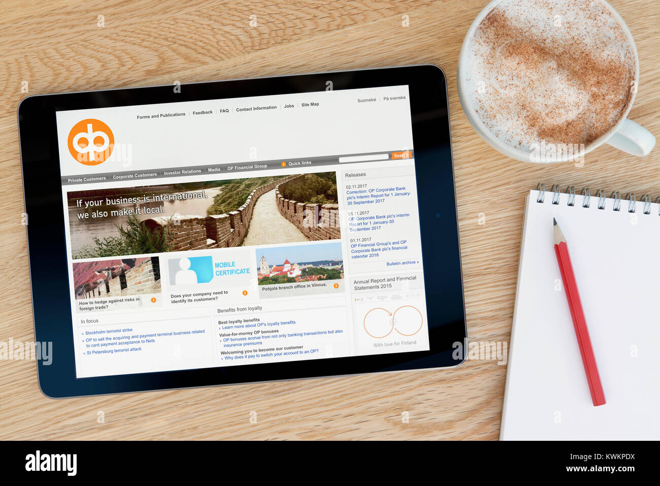 L'OP Financial Group sito web su un iPad dispositivo tablet, appoggiato su un tavolo di legno accanto a un blocco note, matita e tazza di caffè (editoriale solo) Foto Stock