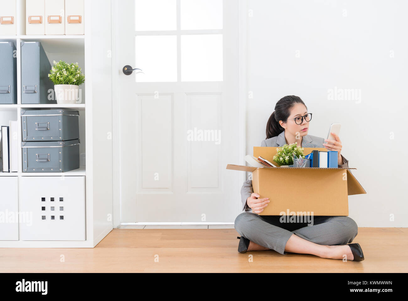 Giovane bella donna d'affari perdendo azienda job seduta sul pavimento di legno in fondo bianco con casella personale e tramite telefono cellulare la ricerca ne Foto Stock