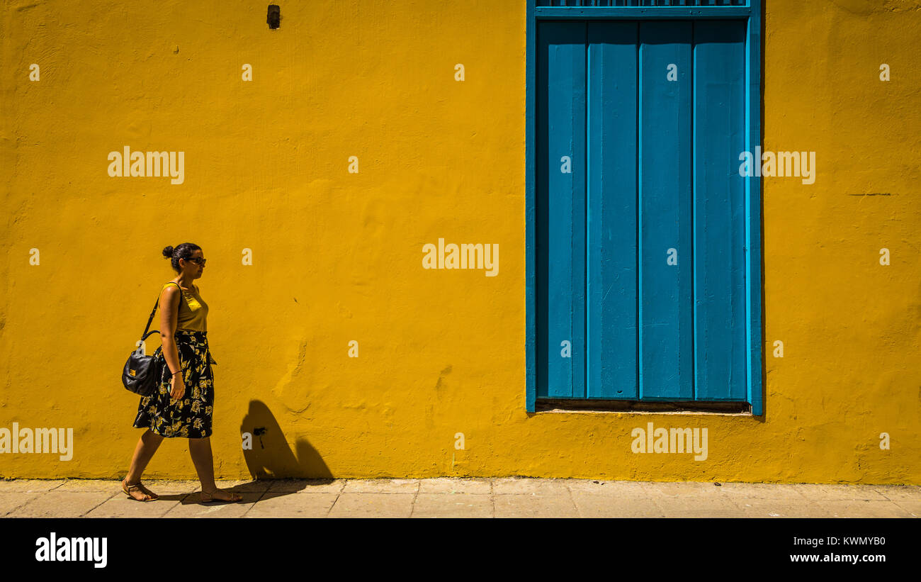 Donna oltrepassando un variopinto edificio giallo con blu brillante di legno chiusa porta o finestra in estate sole su un marciapiede pavimentato Foto Stock