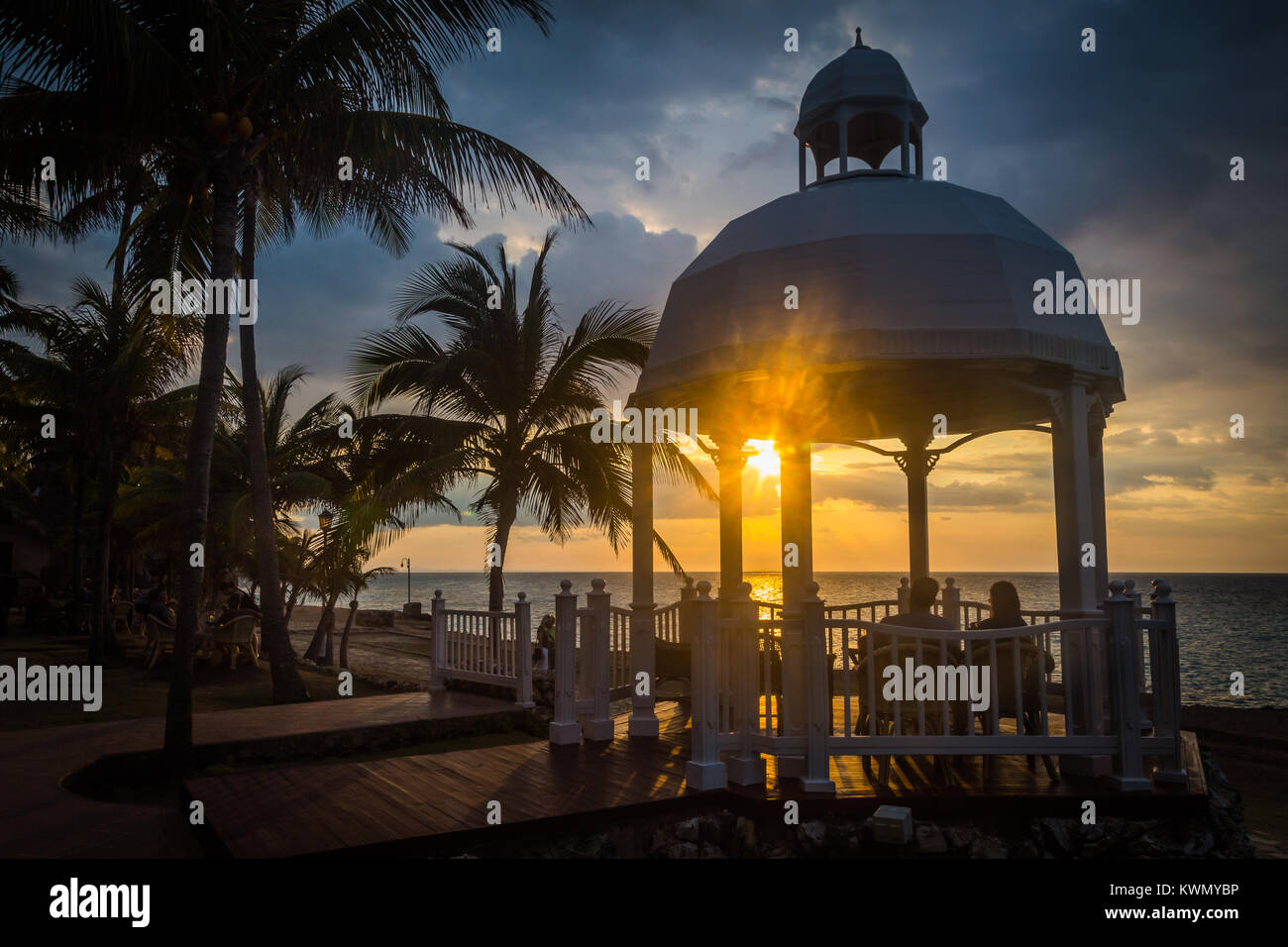 Romantico tramonto tropicale con un giovane seduto vicino insieme su un banco in un gazebo o padiglione con vista sull'oceano con palme lungo il litorale Foto Stock