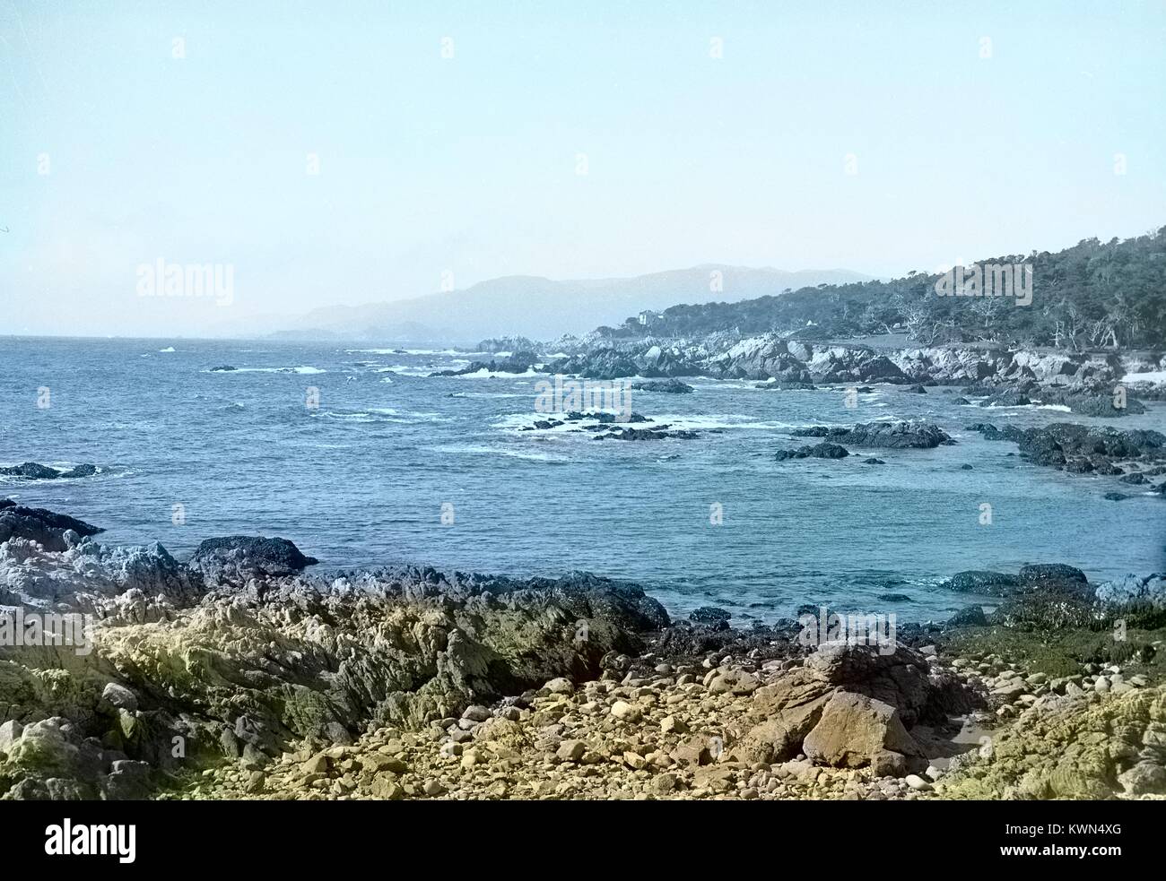 Le onde rompono sulle rocce di origine vulcanica, con scogliere e un piccolo edificio visibile su un punto di terra, sul litorale vicino Monterey, California, 1950. Nota: l'immagine è stato colorizzato digitalmente usando un processo moderno. I colori possono non essere periodo-precisa. Foto Stock