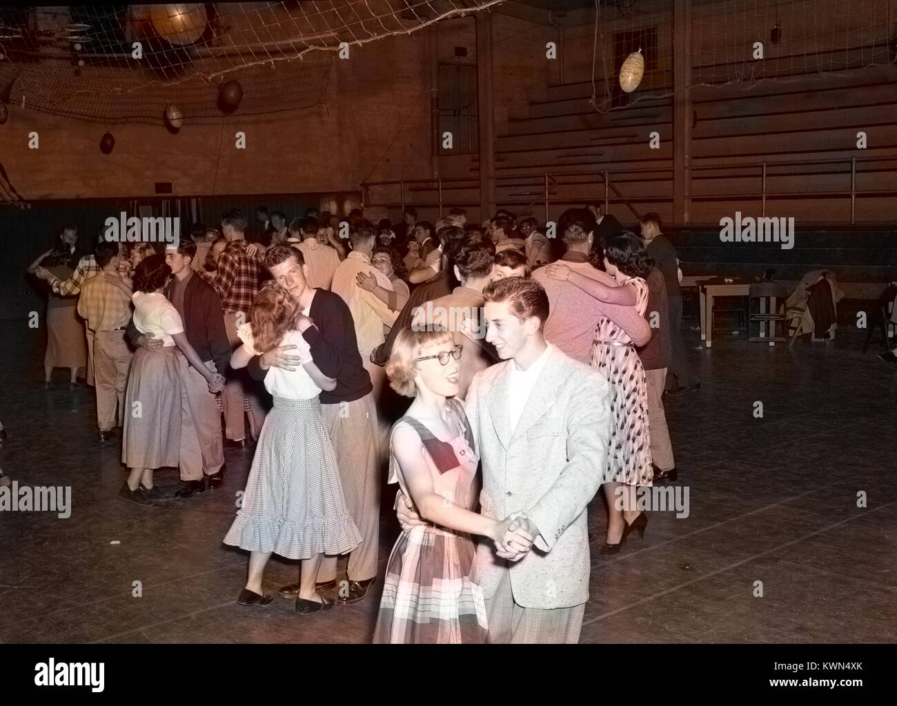 Un giovane uomo e donna giovane tenere mani e ridere insieme mentre slow dancing durante una alta scuola di danza nella palestra a Monterey unione di alta scuola, mentre altre coppie danza in background, Monterey, California, 1950. Nota: l'immagine è stato colorizzato digitalmente usando un processo moderno. I colori possono non essere periodo-precisa. Foto Stock