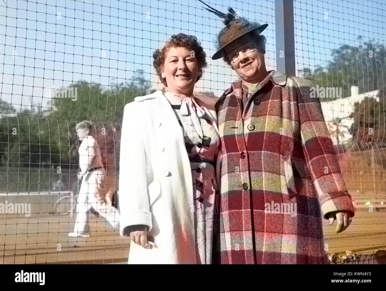 Due donne mature indossando elaborare cappotti, uno con un cappello, stand davanti un campo da tennis e posa per una fotografia, Eureka, California, 1950 Nota: l'immagine è stato colorizzato digitalmente usando un processo moderno. I colori possono non essere periodo-precisa. Foto Stock