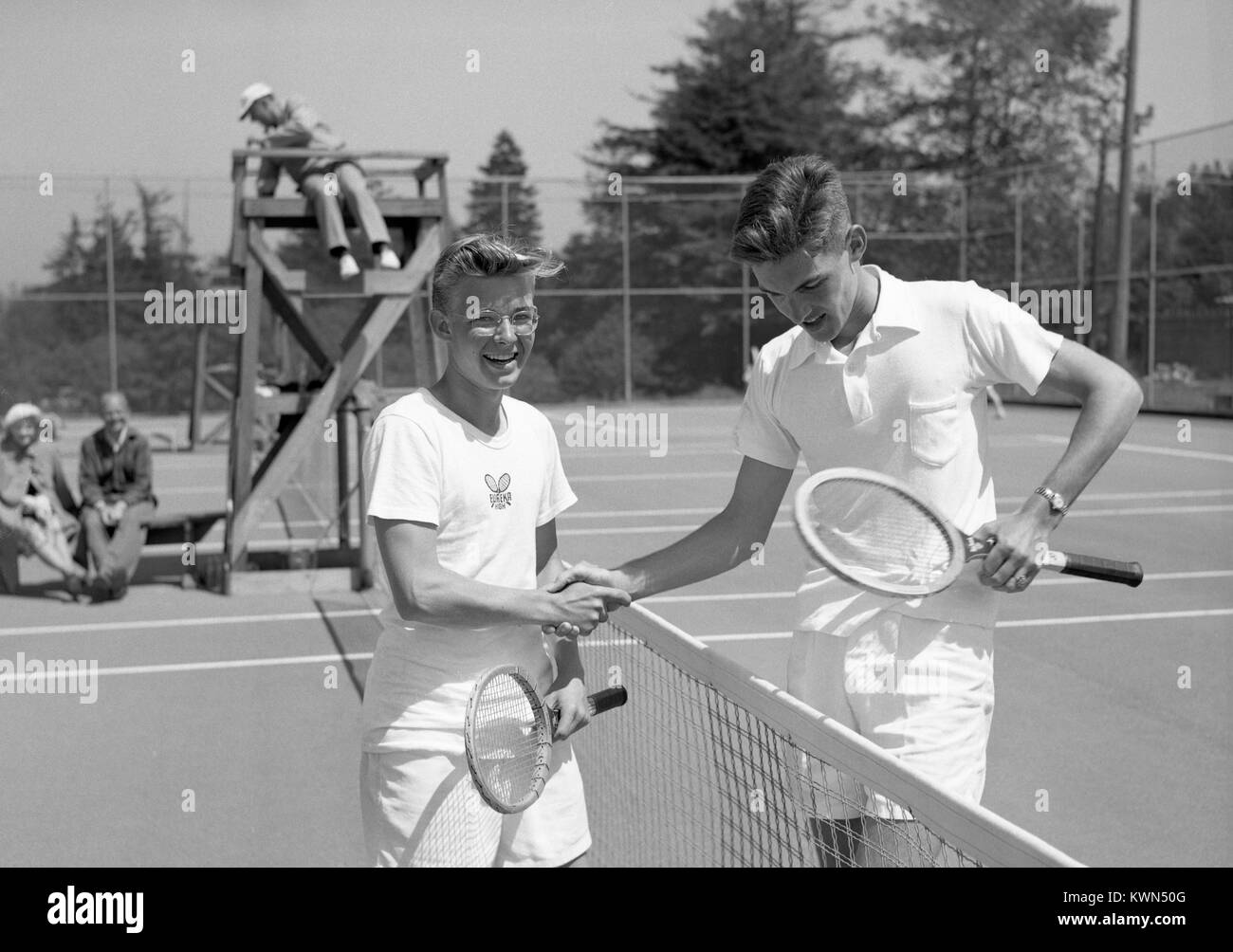 Due giovani ragazzi, uno sostanzialmente più alte rispetto alle altre, agitare le mani sopra la rete di un campo da tennis prima di iniziare una partita con scorekeeper cercando su di un supporto in legno e due adulti maturi osservando dal margine, un ragazzo che indossa un Eureka High School shirt, Eureka, California, 1950. Foto Stock