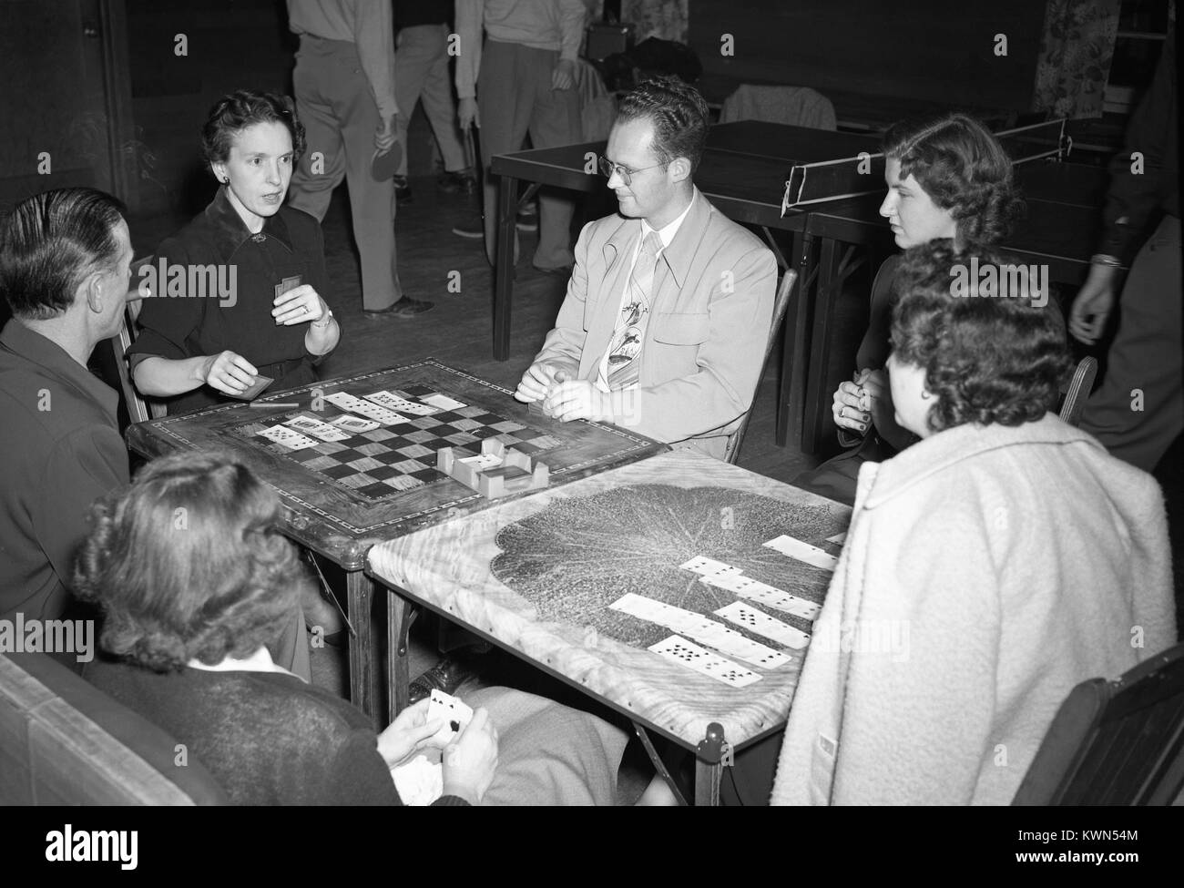 Varie adulti e ragazzi di sedersi attorno a due tavole di carta che sono stati messi insieme e giocare un gioco di carte, con tavolo da ping pong in background, Eureka, California, 1950. Foto Stock