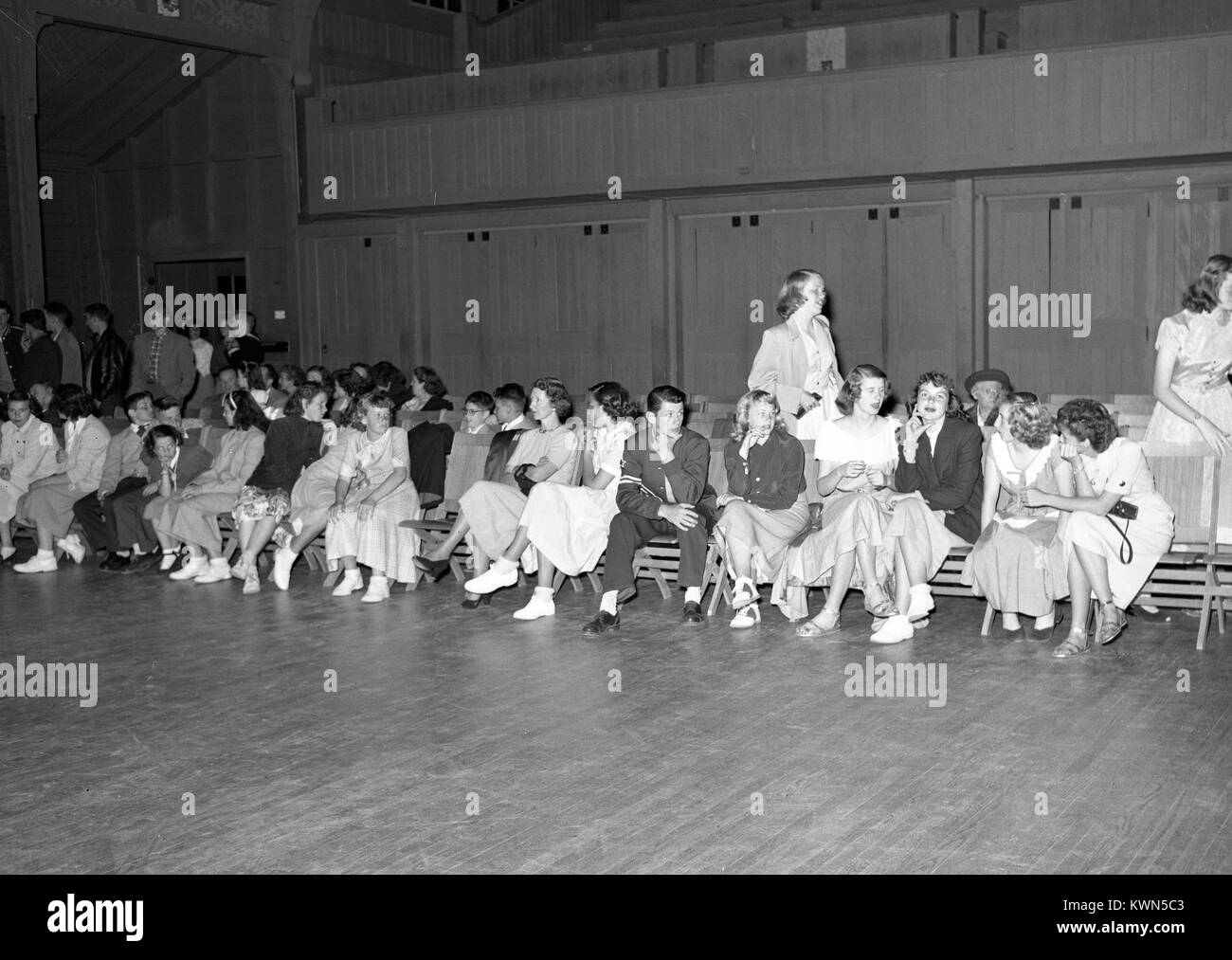 Giovani uomini e donne siedono goffamente insieme su sedie accanto alla pista da ballo, durante una alta scuola di danza nella palestra a Monterey unione di alta scuola, mentre altre coppie danza in background, Monterey, California, 1950. Foto Stock