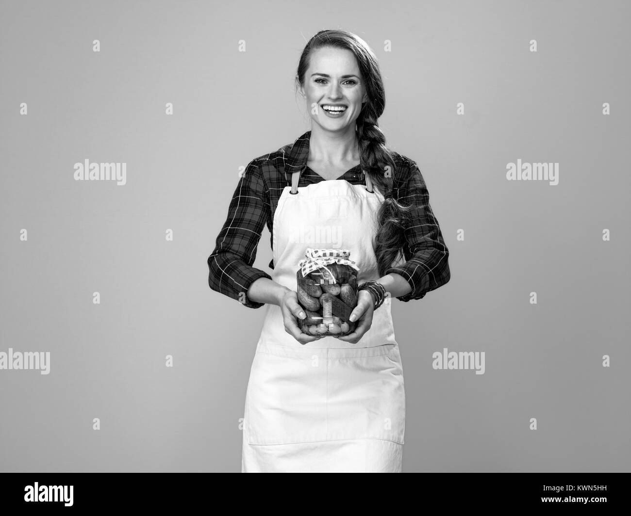 Cibo sano per il vostro tavolo. Ritratto di sorridere donna giovane agricoltore che indossa il grembiule isolato su giallo che mostra un barattolo di cetrioli sottaceto Foto Stock