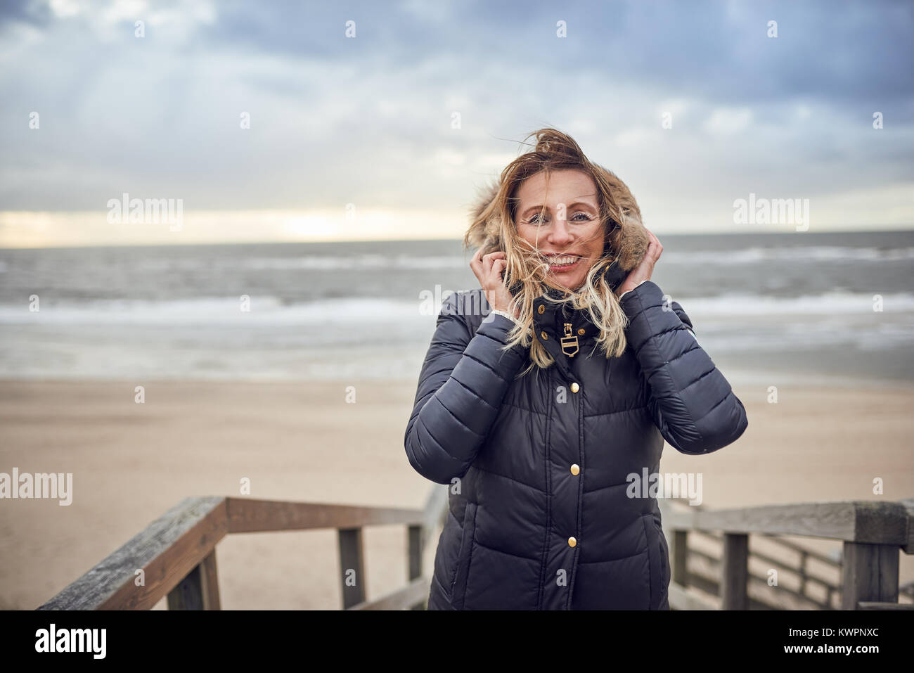 Donna di mezza età sfidando un freddo inverno giornata al mare in piedi su un ponte di legno che si affaccia sulla spiaggia Su breezy day sorridente felicemente in dotazione Foto Stock