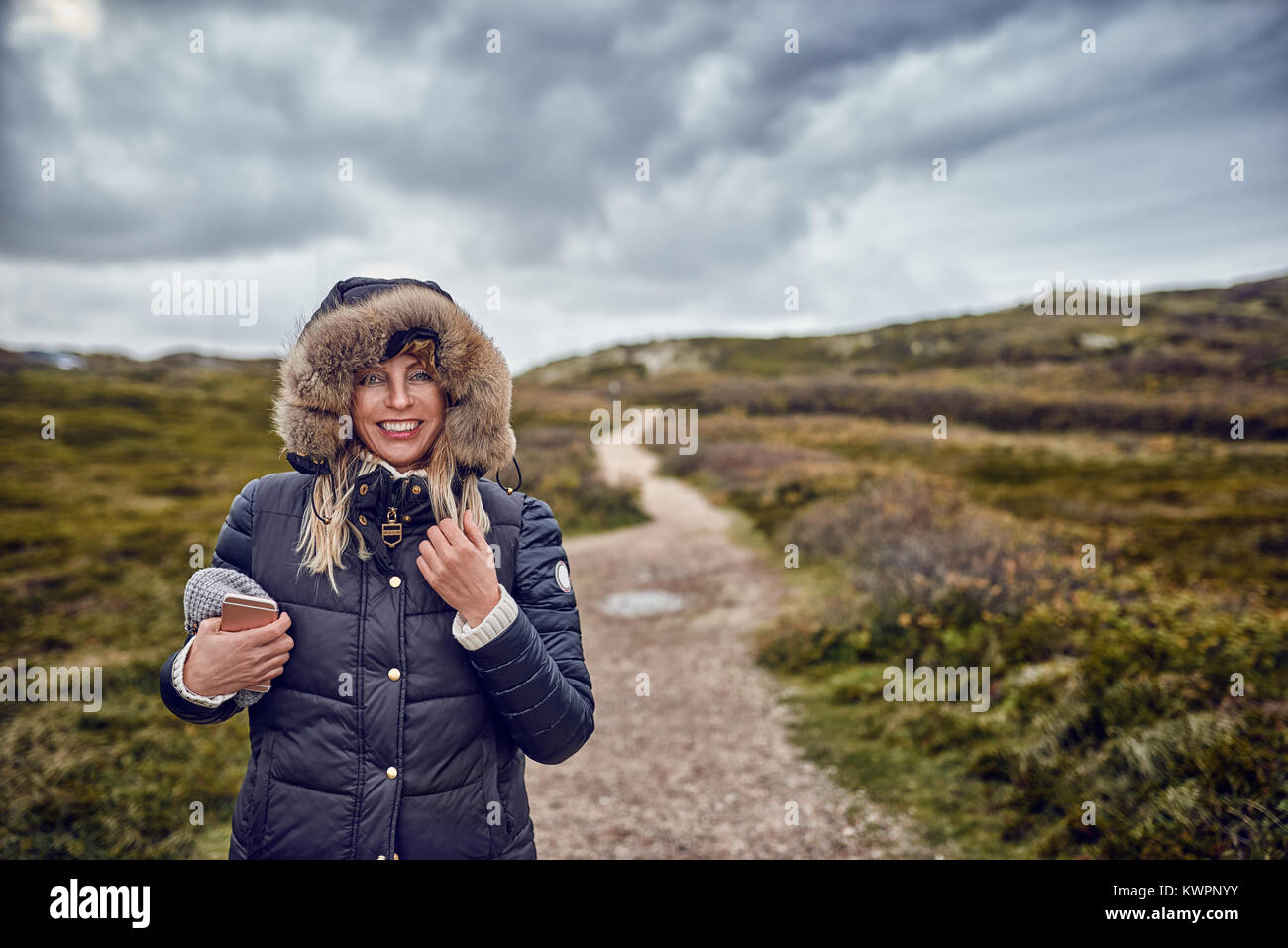 Donna di mezza età sfidando un freddo giorno di inverno in natura a piedi attraverso il paesaggio Su breezy day sorridente felicemente con la videocamera Foto Stock