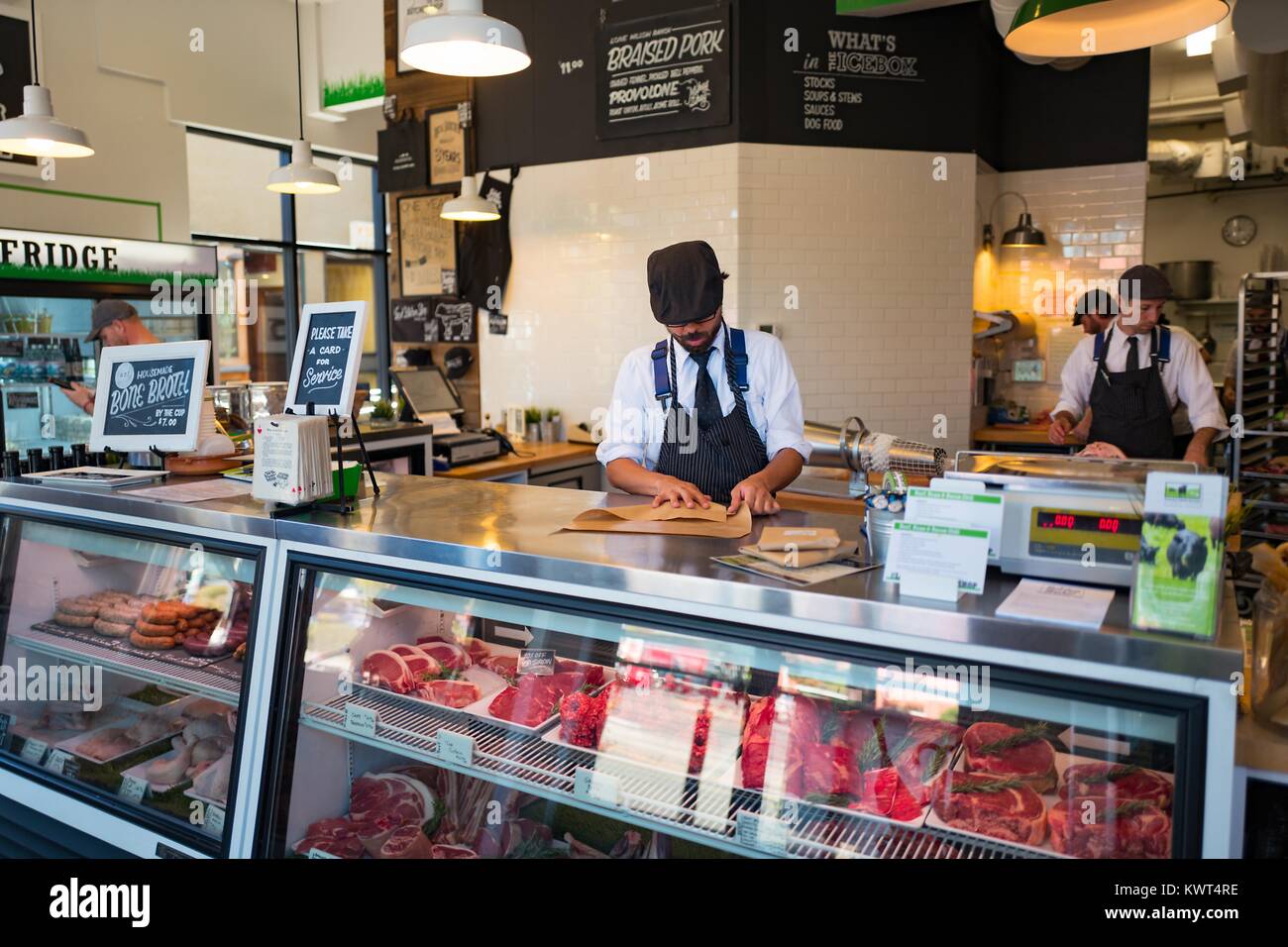 Un uomo che indossa un grembiule a strisce e hat esegue il wrapping di un pezzo di carne, con molti tagli di carne rossa visibile in una vetrina a livello locale macelleria, un artigiano macelleria nel Ghetto Gourmet (Nord Shattuck) quartiere di Berkeley, California, 27 settembre 2017. () Foto Stock