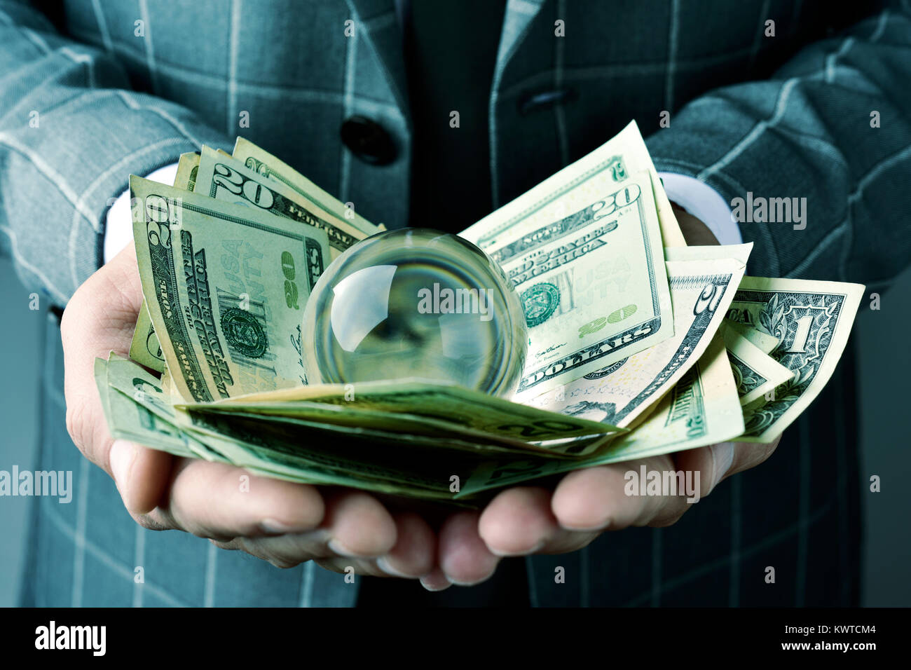 Primo piano di un giovane imprenditore caucasico indossando un elegante abito grigio, con una pila di fatture del dollaro e una sfera di cristallo nelle sue mani Foto Stock