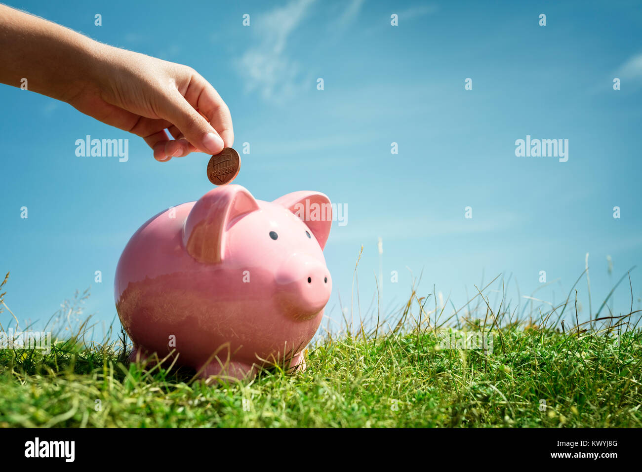 Bambino mano inserire monete e risparmio di denaro nel salvadanaio con erba e cielo blu sullo sfondo Foto Stock