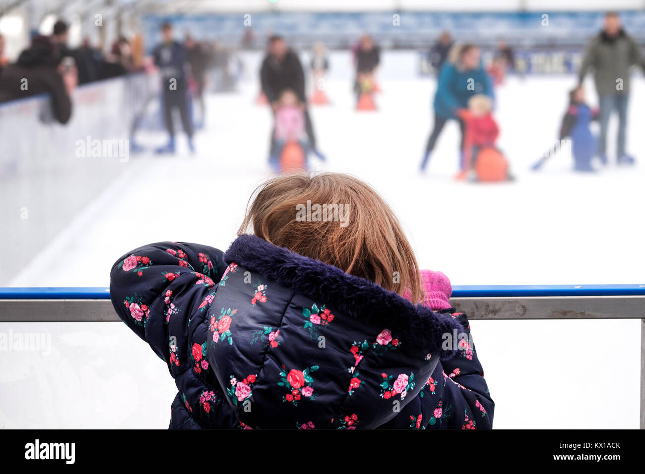 Una giovane ragazza appoggiata sopra la barriera in corrispondenza di una pista per il pattinaggio sul ghiaccio, guardando altre famiglie sul ghiaccio con il pattinaggio aiuti per i bambini. Foto Stock