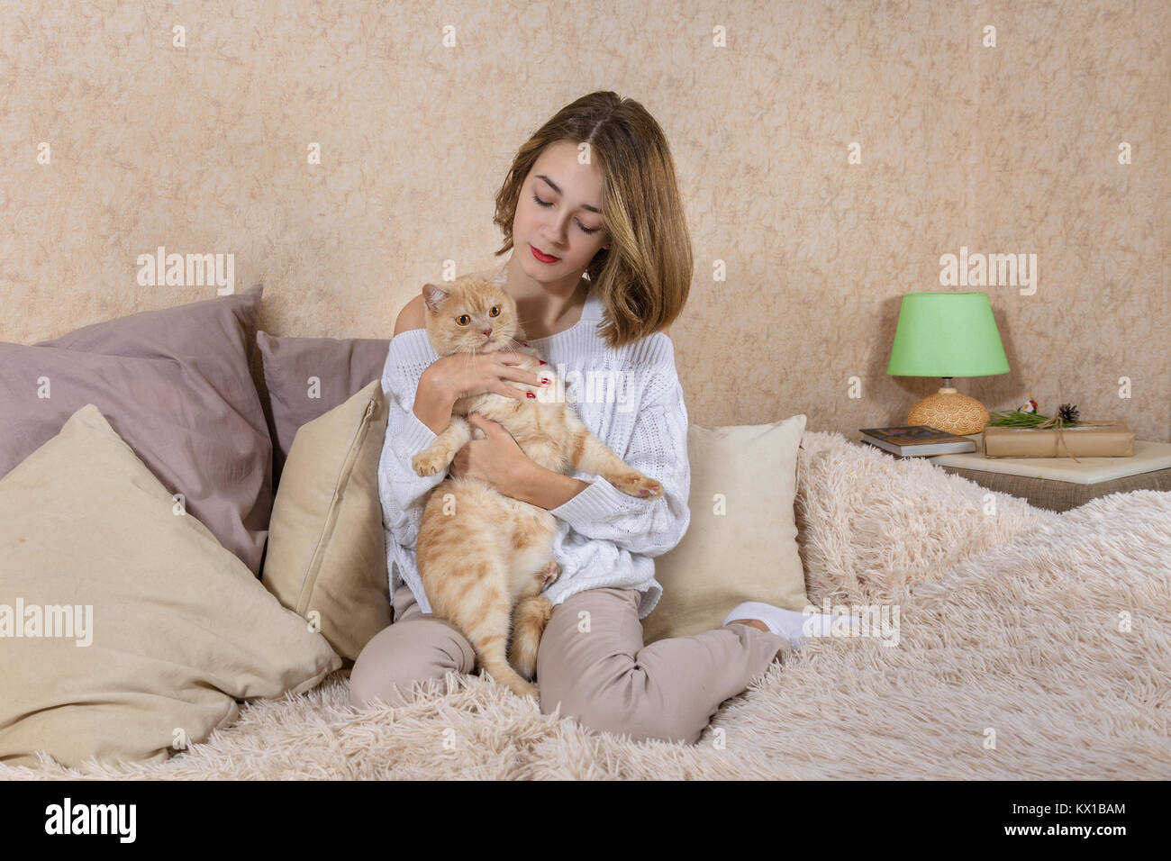 La ragazza in bianco maglione seduta sul divano e abbracciando un gatto rosso Foto Stock