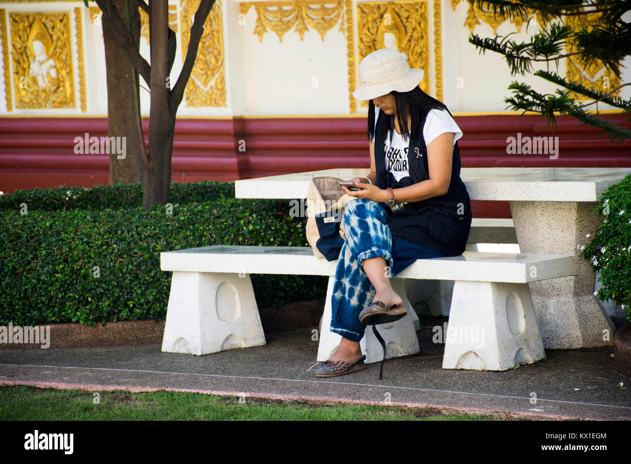 Asian thai donna sedersi e riproduzione di smartphone sul banco di lavoro nel giardino di Wat Phra That Nong Bua tempio di Ubon Ratchathani, Thailandia Foto Stock