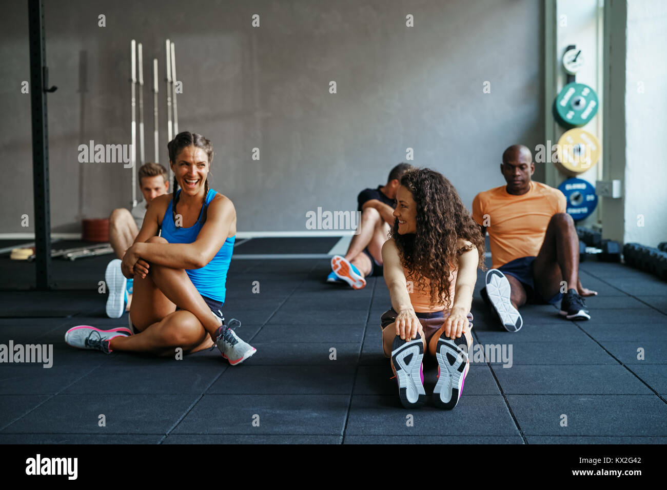 Due donna in abbigliamento sportivo di stretching e di parlare insieme prima di una classe in un club della salute con persone in background Foto Stock