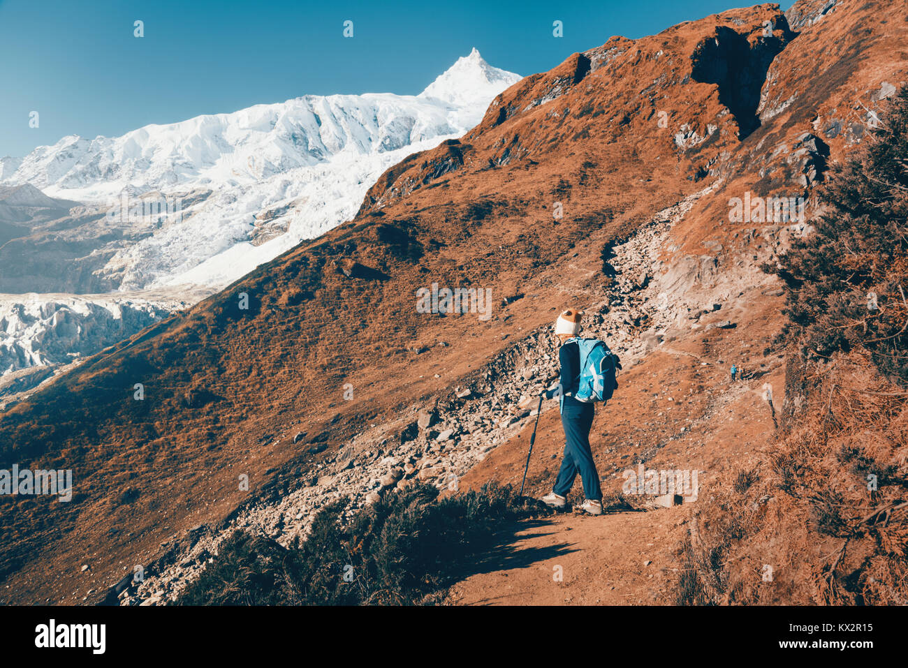 Donna in piedi con zaino sul sentiero di montagna contro la coperta di neve le rocce al tramonto. Paesaggio con ragazza, alte montagne con vette innevate, percorso b Foto Stock