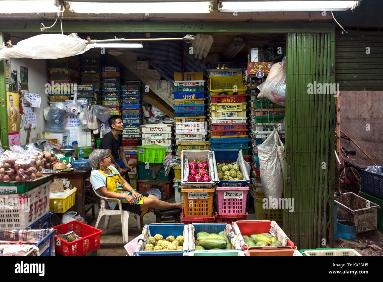 Due frutta asiatica del mercato all'ingrosso titolari di stallo sedersi a loro in stallo a guardare la televisione circondato da frutta esotica. Foto Stock