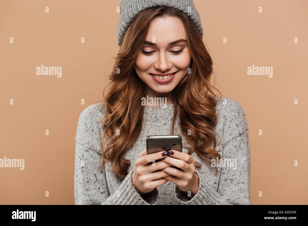 Close-up ritratto di Gorgeous Brunette ragazza nel cappello di lana e un maglione a chattare su smartphone, isolato su sfondo beige Foto Stock