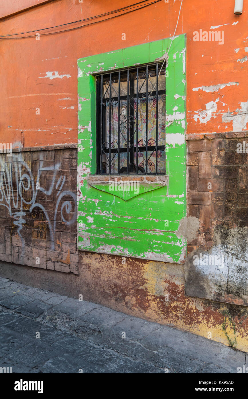 Funky cornice verde intorno ad una doppia finestra, con barre di ferro e un muro di pietra con vernice arancione, alcuni graffiti e una pietra street, in Guanajuato, Messico Foto Stock