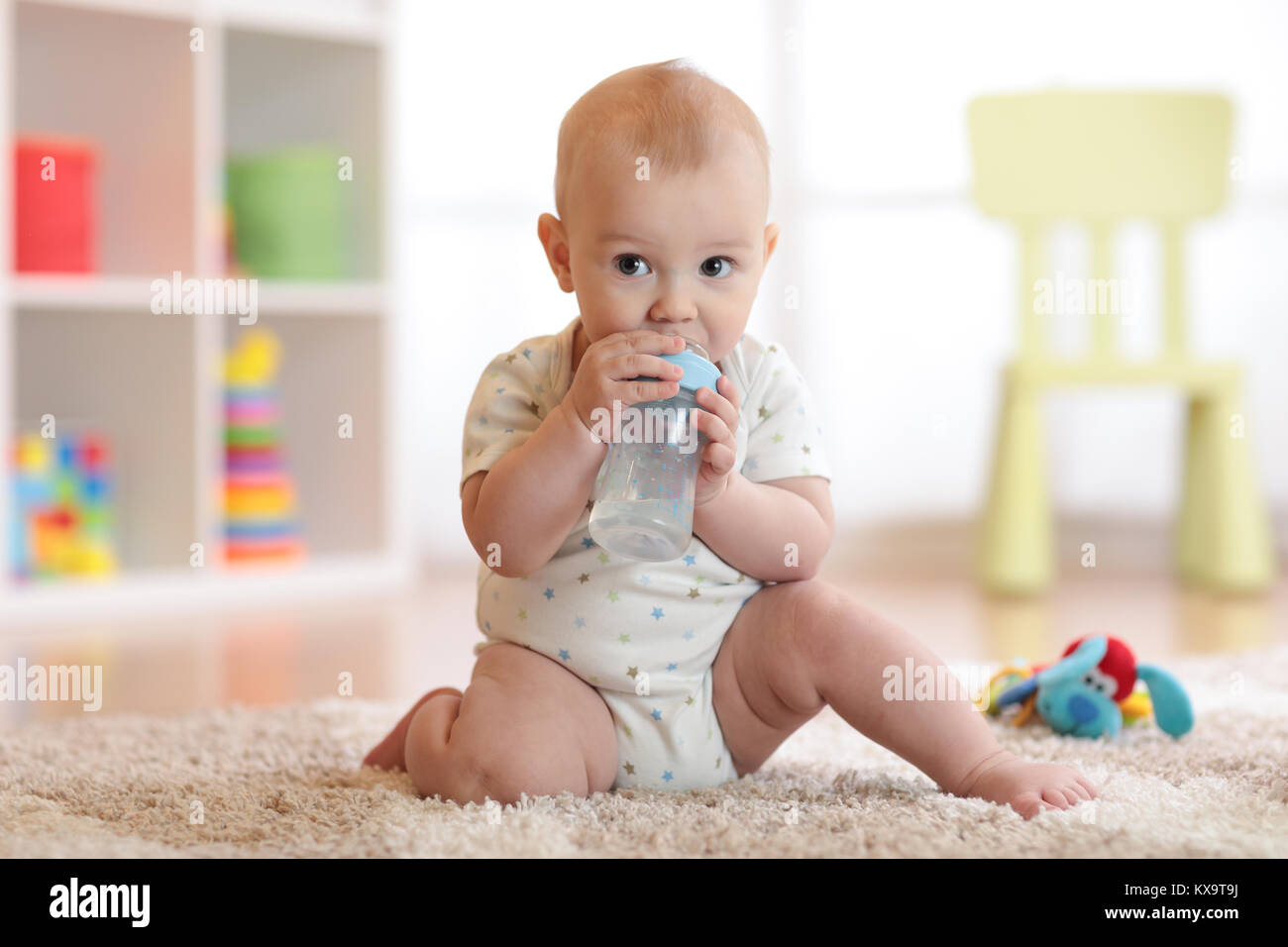 Pretty baby boy di bere dalla bottiglia. Ragazzo seduto su un tappeto o in un asilo nido a casa. Bambino sorridente è di 7 mesi di età. Foto Stock