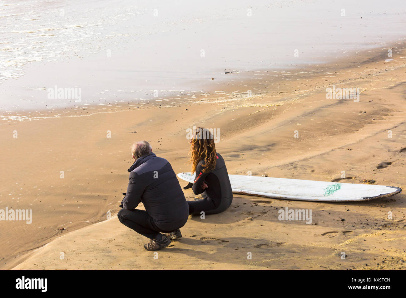 Surfer seduto sulla spiaggia accanto alla tavola da surf con un altro uomo che guarda al mare a Bournemouth Beach, DORSET REGNO UNITO NEL GENNAIO Foto Stock