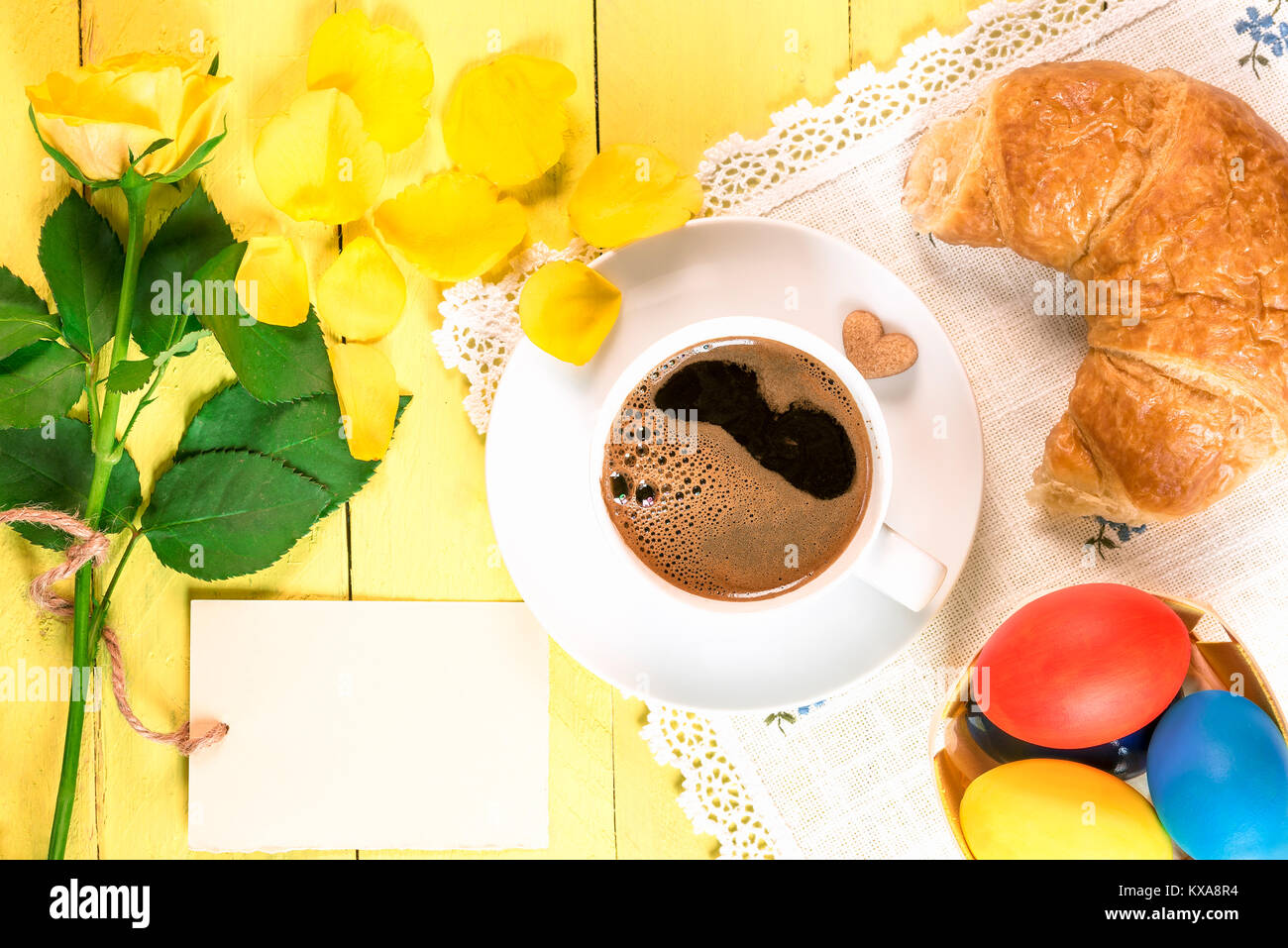 Tavolo per la colazione con una tazza di caffè, pane appena sfornato,  croissant, dipinto di uova di Pasqua e un giallo single rose con petali  caduti e un messaggio di carta Foto