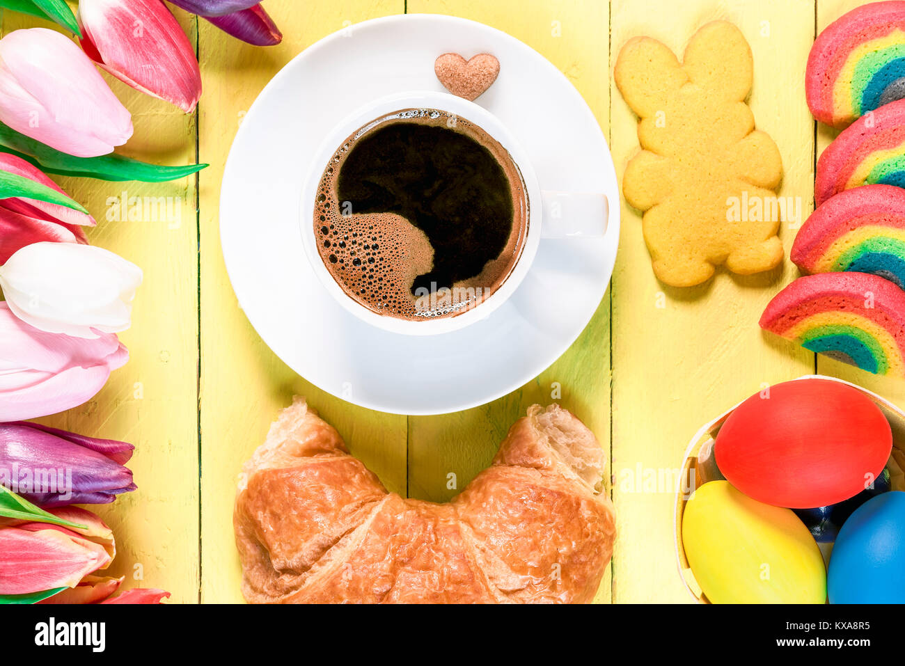 Il mattino di Pasqua con una tazza di caffè, rainbow cookies, dipinto di uova, bunny cookie sagomato, croissant e tulipani multicolore, su un giallo tavolo in legno. Foto Stock