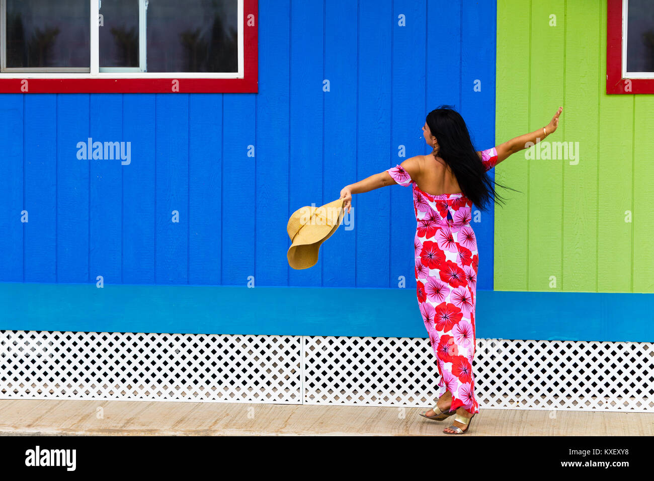 Piuttosto Polynesian islander in un rosso vestito floreale e cappello ballando nella parte anteriore di un colorato di verde e blu della parete in legno in Hawaii Foto Stock