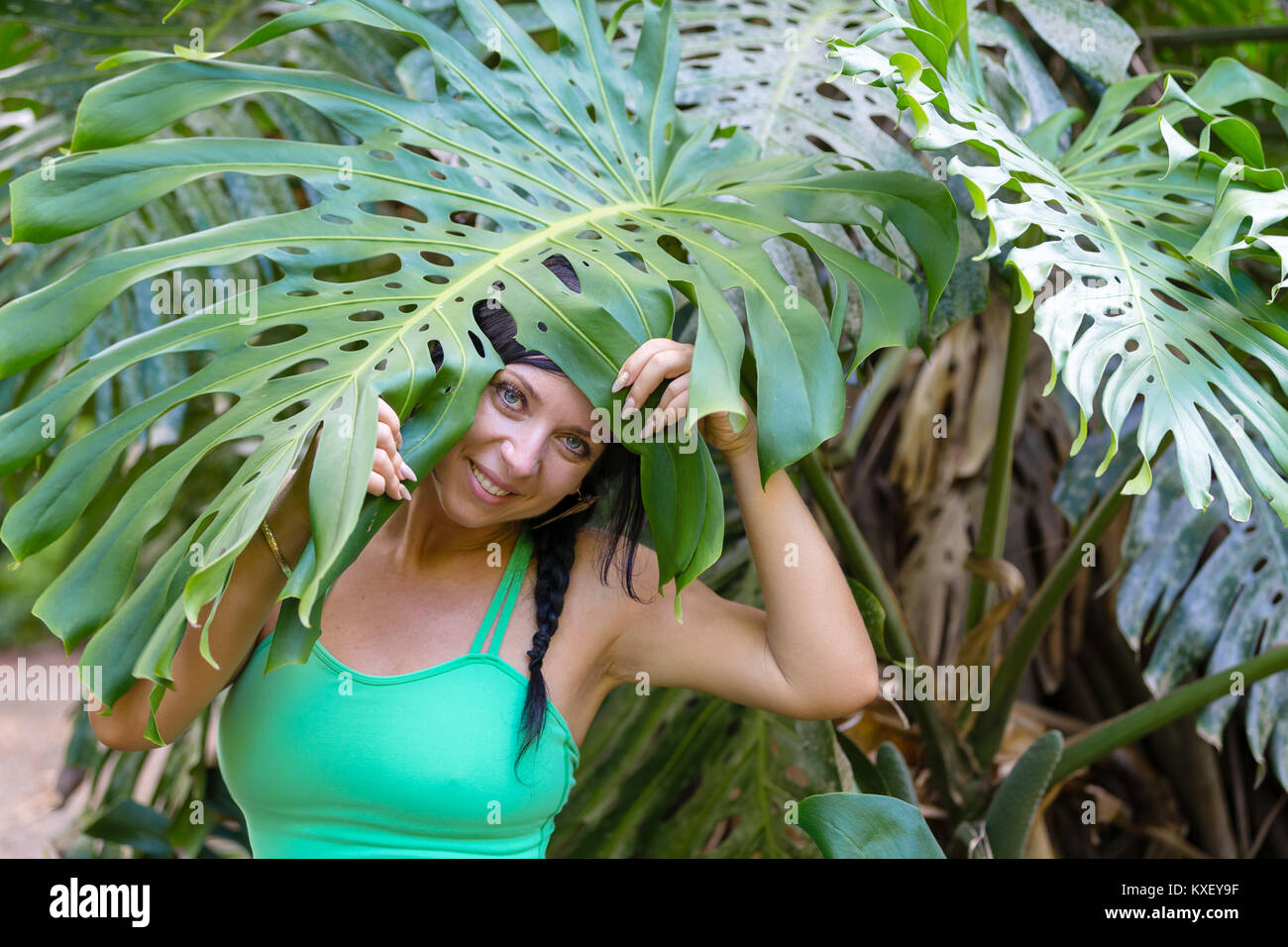 Carino donna giocoso che spuntavano da sotto una grande foglia verde di monstera deliciosa, o Delicious Monster, impianti su Oahu, Hawaii con un sorriso Foto Stock