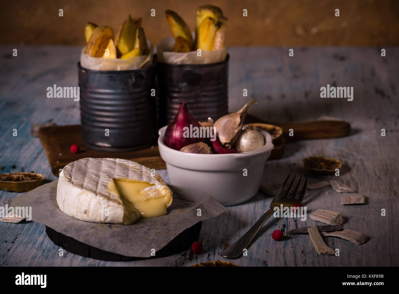 La foto in orizzontale del fuso a caldo di formaggio camembert sul foglio di carta con ciotola piena di aglio e cipolle e le vecchie lattine vintage con patate fritte le strisce sulla bl Foto Stock