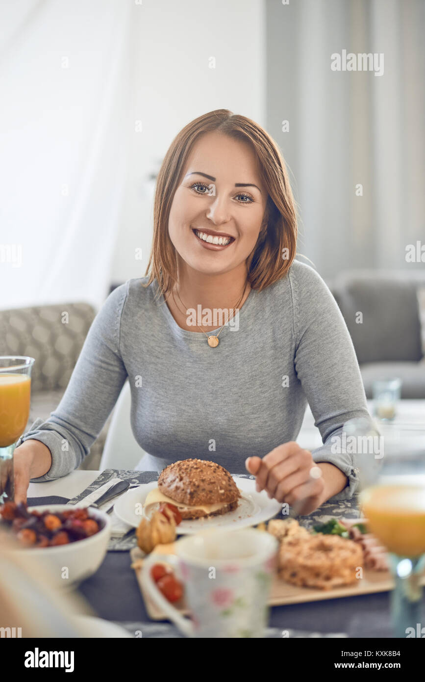 Felice donna attraente godendo un sandwich per colazione seduti ad un tavolo apparecchiato con cibo sorridente verso la telecamera Foto Stock