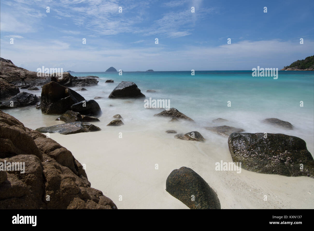 Una lunga esposizione delle onde di luce di laminazione su rocce di grandi dimensioni in un idilic spiaggia di sabbia bianca in una piccola baia con piccole isole all'orizzonte. Foto Stock