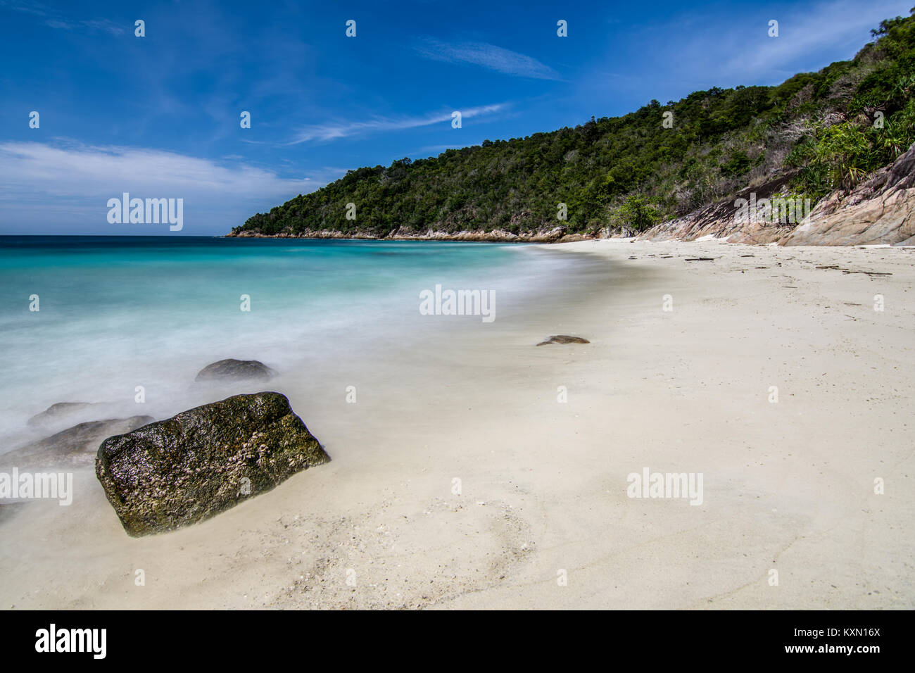 Una lunga esposizione delle onde dal tranquillo sul mare tropicale di spiaggia di sabbia bianca con struttura ad albero collina coperta e lo sfondo blu del cielo. Foto Stock