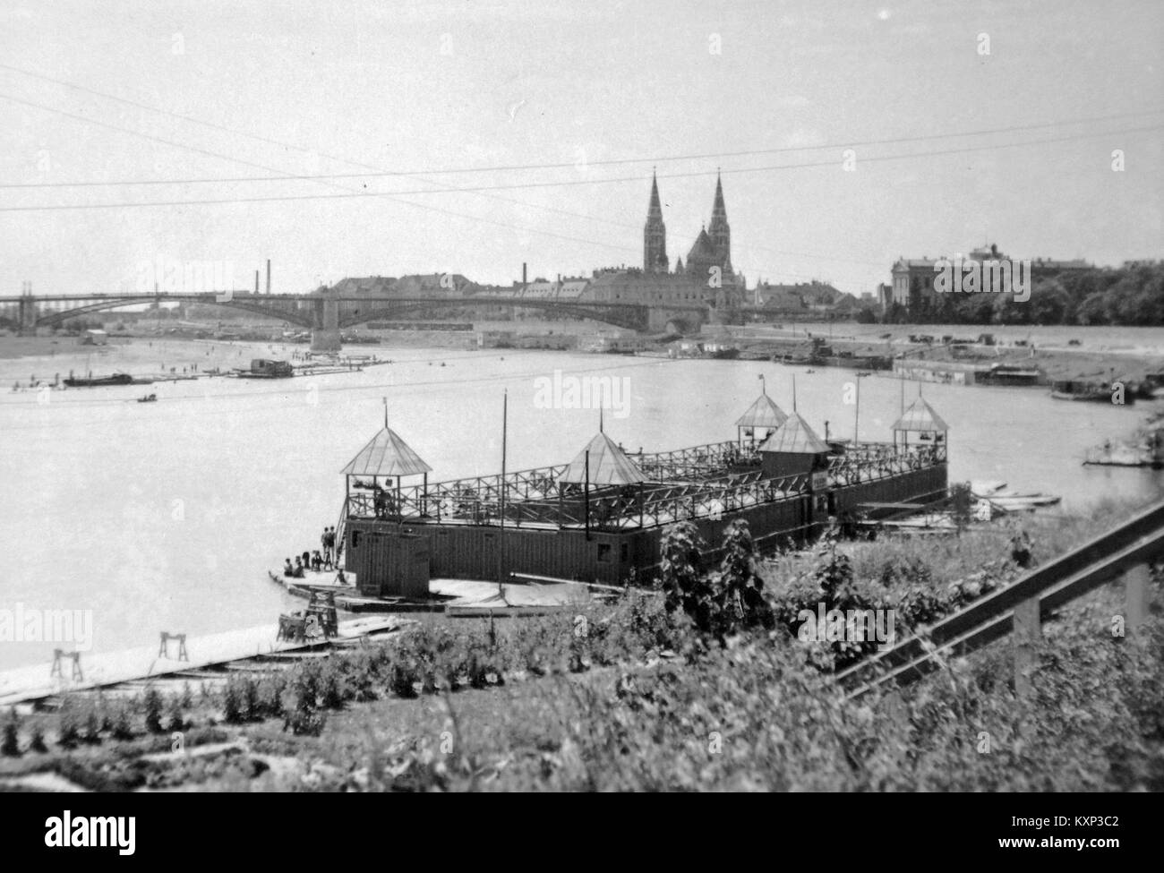 Una fotografia storica che mostra un bagno sul fiume Tisza in Ungheria, con il vecchio ponte stradale e la chiesa di voto sullo sfondo. L'immagine cattura un momento della vita ungherese all'inizio del XX secolo. Foto Stock