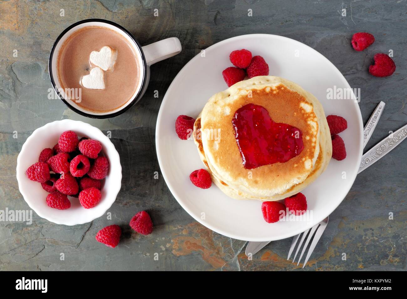 Frittelle con marmellata a forma di cuore, cioccolato e lamponi su un sfondo di ardesia. Il concetto di amore. Foto Stock