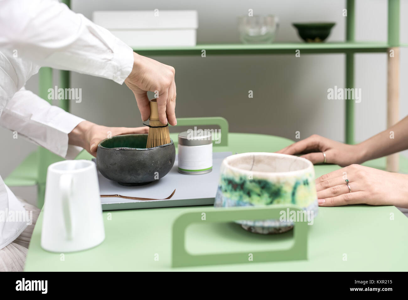 Le donne preparano un cinese tè verde Matcha sul verde del tavolo in metallo per interni. Una ragazza che mescola il tè con una frusta nel recipiente scuro. Sul tavolo c'è un Foto Stock