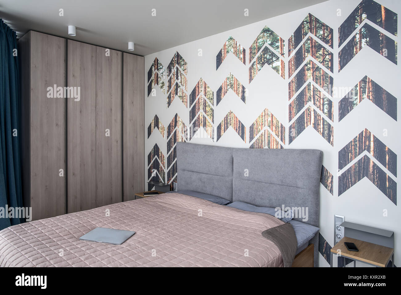 Camera da letto in stile moderno con una parete colorata con stampe di pino. Vi è un letto con una coperta e cuscini e un laptop, comodini con libro e ce Foto Stock