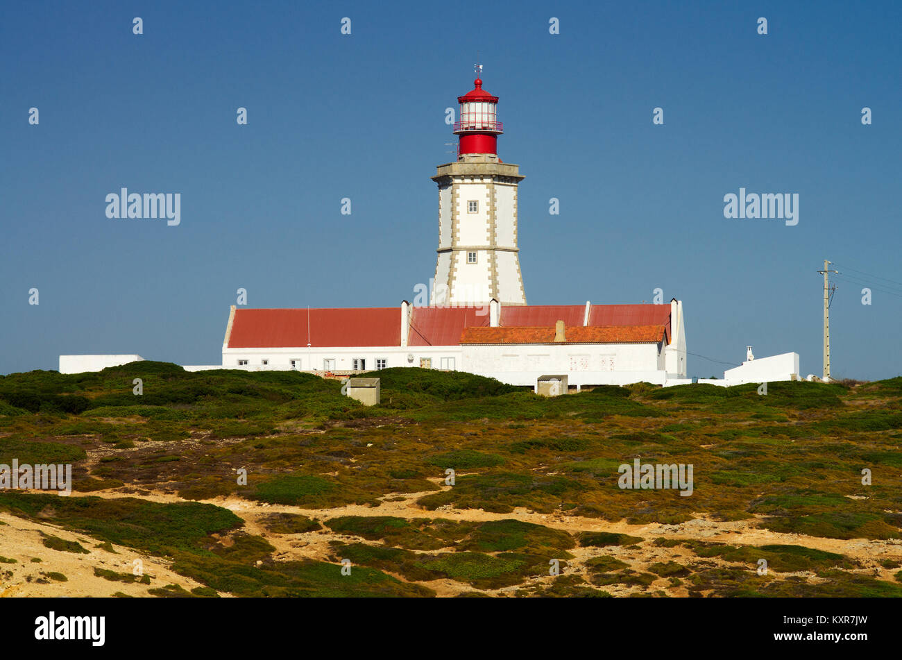 Cape Espichel faro come visto dal lato dell'oceano. Bassa vegetazione, pareti di colore bianco e rosso e rabboccato faro torre contro il cielo blu. Sesimbra, Foto Stock