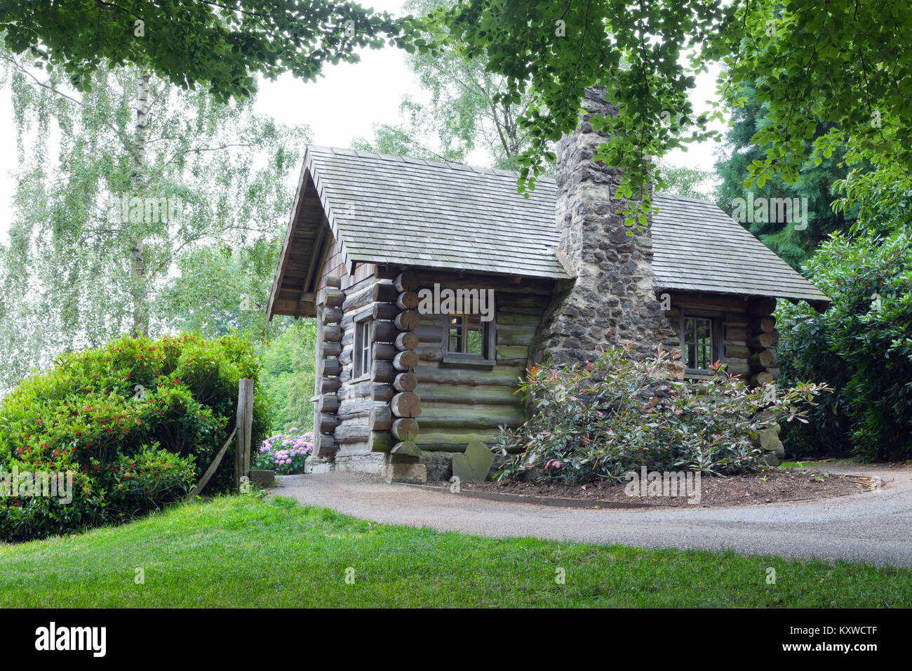 Vecchio cottage di legno con pietra camino esterno nel giardino fiorito con alberi maturi . Foto Stock