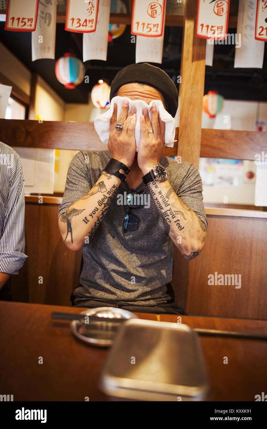 Uomo con bracci tatuati seduti ad un tavolo in un ristorante, tergi il suo volto con il panno umido. Foto Stock