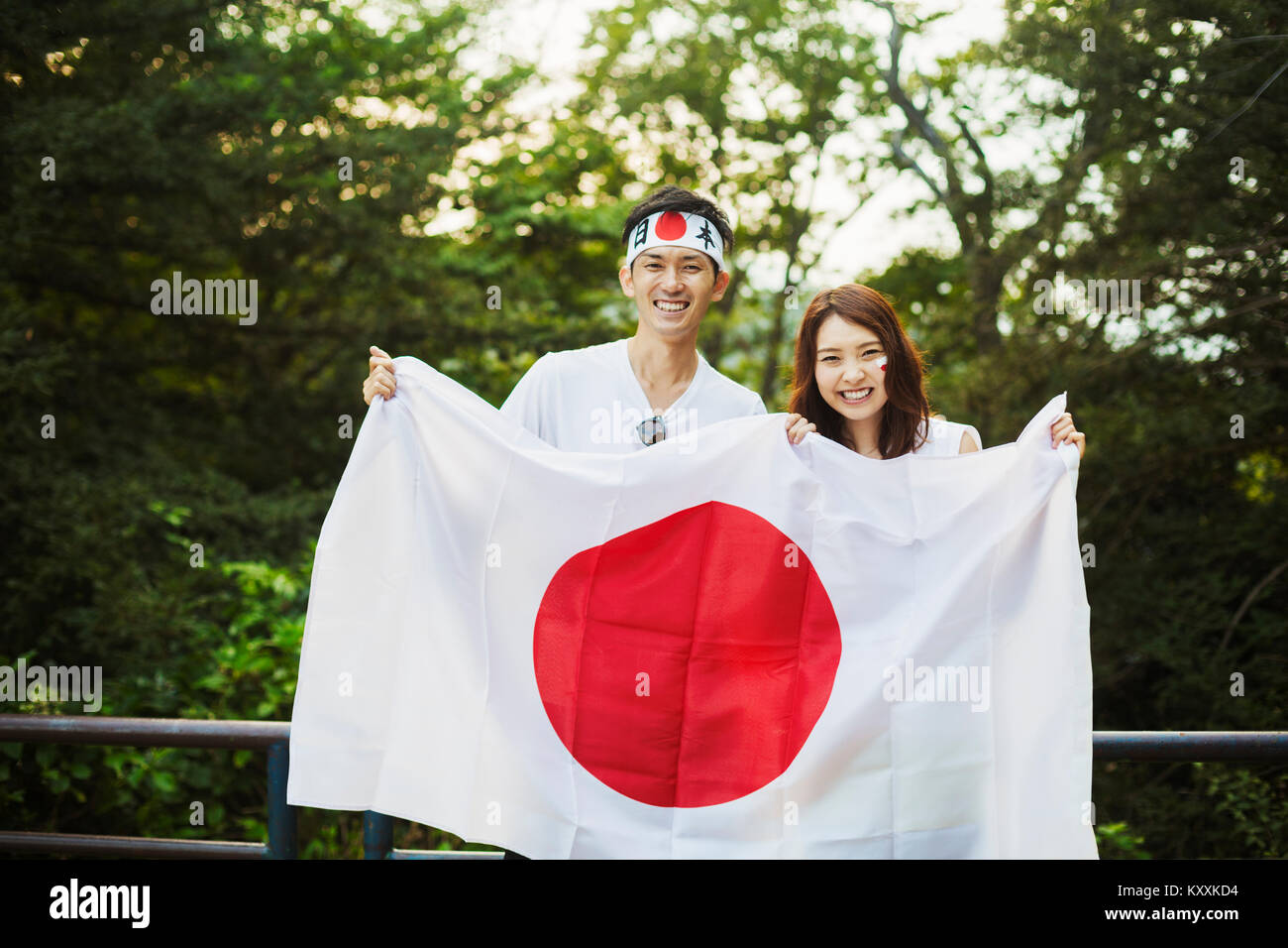 Uomo che indossa la fascia e la giovane donna con capelli castani e faccia tenuta vernice bandiera giapponese, sorridente alla fotocamera. Foto Stock