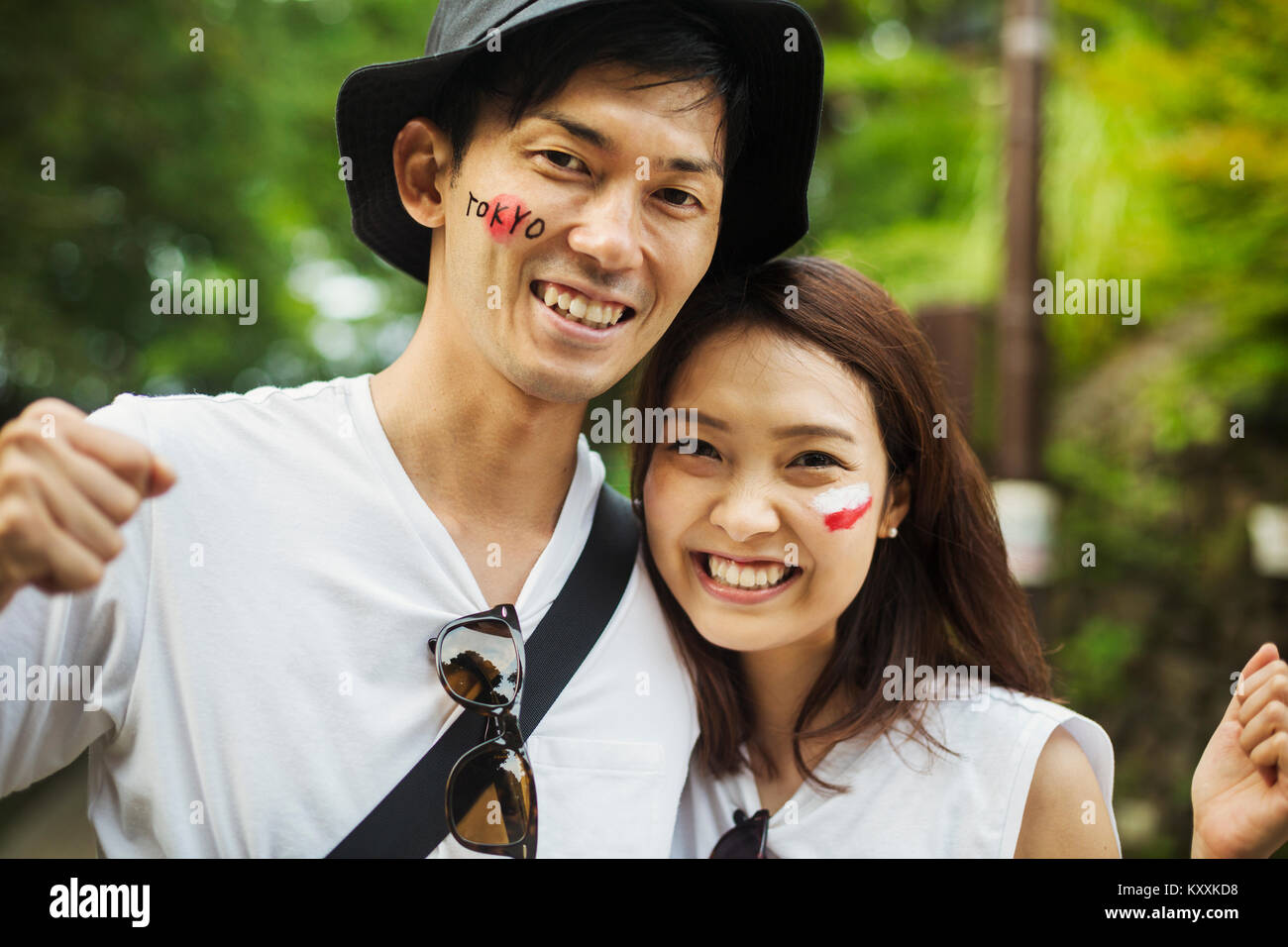 Ritratto di uomo che indossa un cappello e giovane donna con capelli castani, bandiera giapponese dipinta sulla sua guancia, sorridente alla fotocamera. Foto Stock