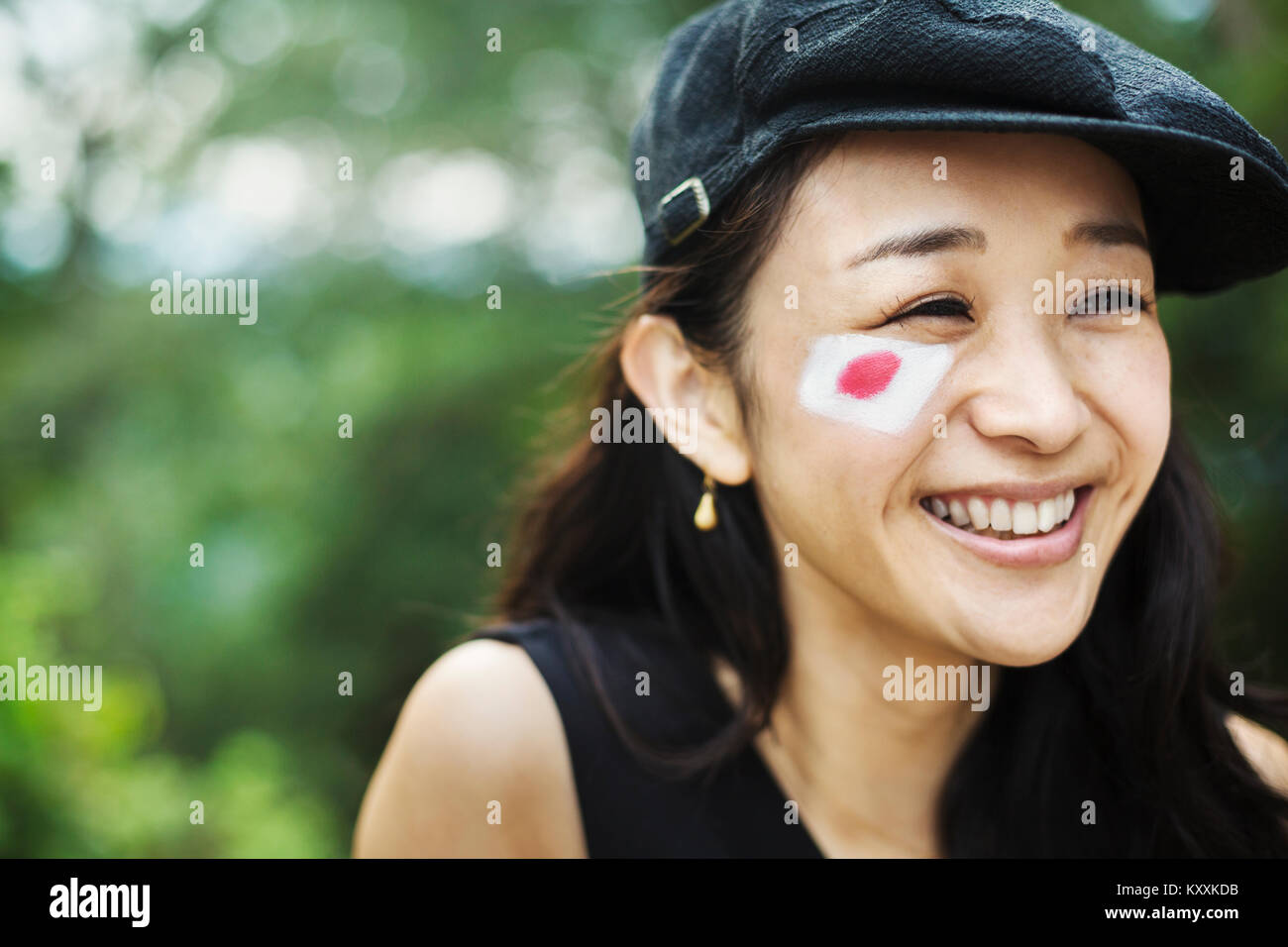 Ritratto di sorridente giovane donna con i capelli neri che indossa il tappo piatto, bandiera giapponese dipinta sulla sua guancia. Foto Stock